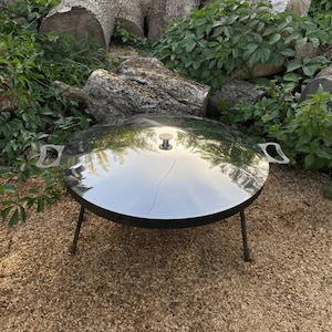 May include: A large, round, stainless steel outdoor cooking surface with a reflective finish. It has a small, round handle on top and two side handles. The cooking surface is supported by three black metal legs, suitable for outdoor grilling.
