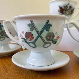 Op de afbeelding: Set van drie witte theekopjes met schoteltjes. Elk kopje heeft een ander ontwerp. Een kopje heeft een ontwerp van twee gekruiste sleutels over een roos en een dobbelsteen met een oog.