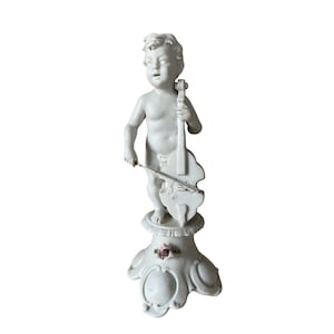 Pode incluir: Figurine de porcelana branca de um querubim tocando um violoncelo, em pé sobre um pedestal com uma rosa rosa.