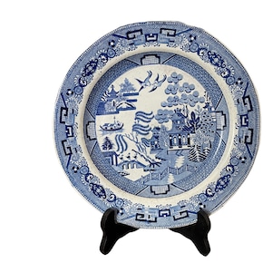 May include: A blue and white porcelain plate with a traditional Chinese design. The plate features a landscape scene with a pagoda, trees, and a river. The border of the plate is decorated with a repeating pattern of geometric shapes.