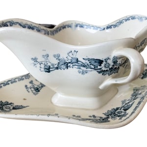 Może przedstawiać: Biała porcelanowa sosjerka z niebieskim wzorem kwiatowym i pasującą podstawką. Sosjerka ma uchwyt i falisty brzeg.