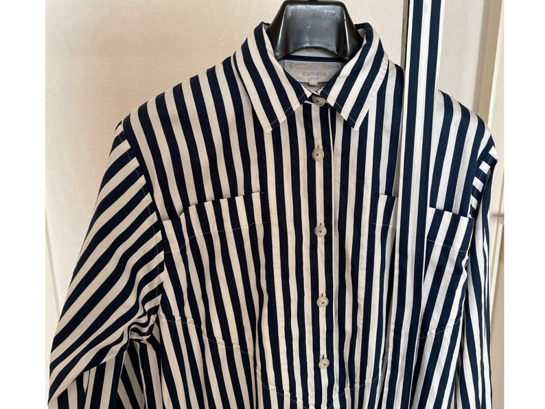 May include: A long-sleeved, striped shirt with a white and navy blue pattern. The shirt has a button-up front and a collar. The shirt has two pockets on the front.