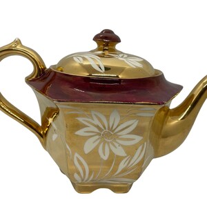 Könnte beinhalten: Eine sechseckige Teekanne mit metallisch goldener Oberfläche und weißen Blumenmustern. Der obere Rand und der Deckel sind in einem tiefen Bordeauxrot abgesetzt. Sie verfügt über einen geschwungenen Henkel, einen passenden Deckel und einen Ausguss in Gold.