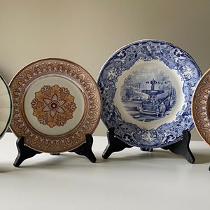 Könnte beinhalten: Vier antike Keramikteller mit dekorativen Designs. Die Teller werden auf schwarzen Ständern präsentiert. Ein Teller zeigt ein blaues und weißes Design mit einer Fontänenszene. Zwei Teller haben ein braunes und goldenes Design mit einem floralen Motiv. Der vierte Teller hat ein braunes und weißes Design mit einer Szene von Menschen auf einem Markt.