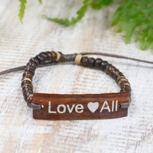 Peut inclure: Un bracelet en bois brun avec des perles brunes et une étiquette en bois qui dit "Love All" avec un symbole de cœur.