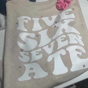 May include: A beige sweatshirt with the words "FIVE SIX SEVEN ATE" printed in white in a retro font. The letters are outlined in a light pink color.