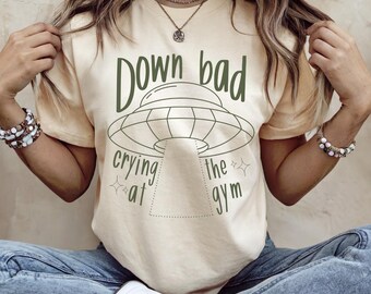 May include: Cream-colored t-shirt with the text "Down bad crying at the gym" in dark green, featuring a UFO graphic. The shirt is a casual style.