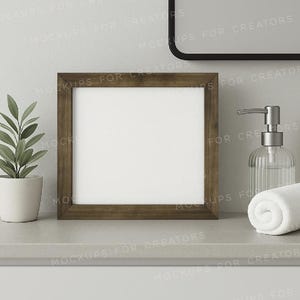May include: A square, brown-framed picture frame with a white interior sits on a gray surface. A small potted plant, a clear soap dispenser, and a rolled white towel are also on the surface. A black-framed mirror is in the background.