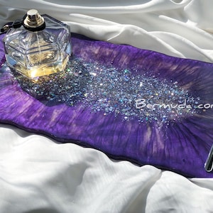 Puede incluir: Bandeja de servir de resina púrpura con un asa plateada y purpurina iridiscente. La bandeja tiene un borde ondulado y está decorada con un patrón de remolino. Una botella de perfume de vidrio transparente está sentada en la bandeja.