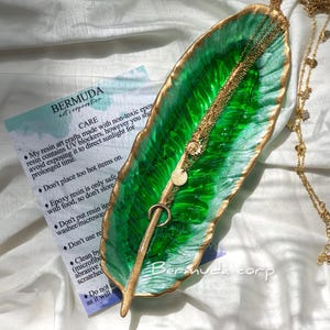May include: A green and gold resin leaf tray with a gold rim. The tray is decorated with a gold leaf design and has a smooth, glossy finish. The tray is perfect for holding jewelry, keys, or other small items.