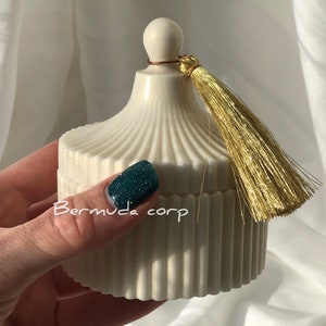 Könnte beinhalten: Weiße Kerzenvase aus Keramik mit goldener Quaste und geripptem Design. Die Vase wird in einer Hand mit grünem Nagellack gehalten.