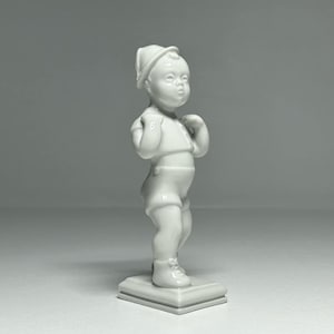 Puede incluir: Figurilla de porcelana blanca de un niño con un sombrero de pie sobre una base cuadrada.