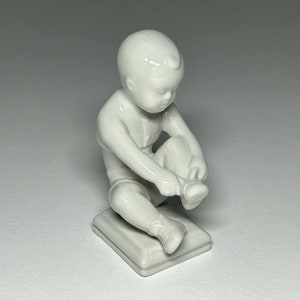 Puede incluir: Una figurita de porcelana blanca de un niño sentado y atándose el zapato.