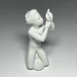 Puede incluir: Una figurita de porcelana blanca de una figura arrodillada sosteniendo un pájaro en sus manos.