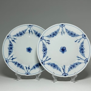 May include: Two white porcelain plates with blue floral designs. The plates have a raised, textured surface and are displayed on clear plastic stands.