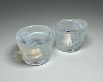 Puede incluir: Dos vasos de chupito de vidrio transparente con un diseño de remolino blanco. Los vasos tienen el texto "Kosta Boda" impreso en la parte inferior.