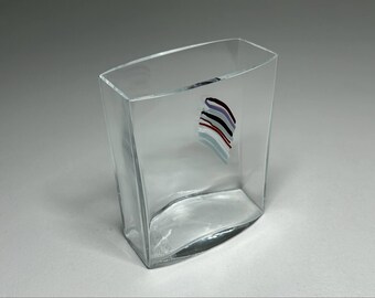 May include: A clear, rectangular glass vase with a curved base and a colorful abstract design on the front. The design features a series of curved lines in red, black, white, and blue. The vase has a modern, minimalist aesthetic.