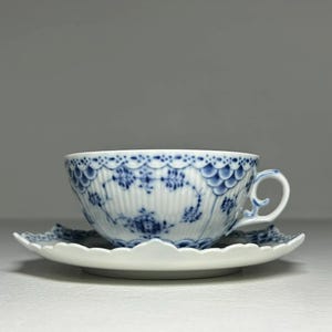 May include: A white teacup and saucer set with blue floral and scalloped trim details. The teacup has a curved handle and a delicate, detailed design. The saucer has a scalloped edge, complementing the cup's design.