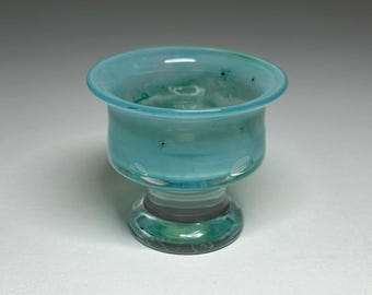 Puede incluir: Un pequeño cuenco de vidrio hecho a mano, de color azul turquesa. El cuenco tiene un borde redondeado y una base de pedestal. Se ven diseños decorativos recortados cerca de la base. El vidrio es translúcido, permitiendo el paso de la luz. Un objeto decorativo.