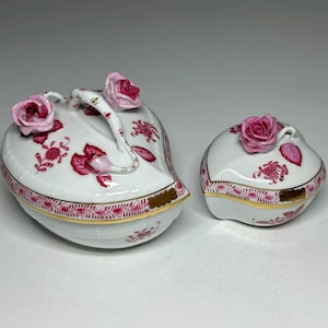 May include: Two white porcelain trinket boxes with pink floral designs and gold trim. One box is heart-shaped with a handle and rose accents. The other is a smaller, rounded box with a rose on top. Decorative and elegant.