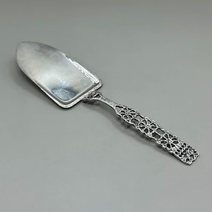 May include: A silver cake server with a wide, rectangular blade and a decorative, openwork handle. The handle features a floral design. The server is made of a shiny, reflective metal, likely stainless steel or silver.