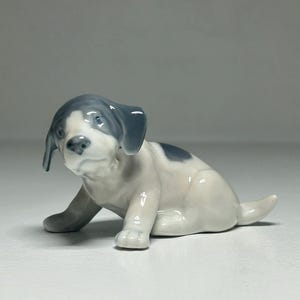Puede incluir: Una figura de cachorro de cerámica en posición sentada. El cachorro tiene un cuerpo blanco con manchas grises en la cabeza y la espalda. La figura es brillante y detallada, mostrando una expresión juguetona e inocente.