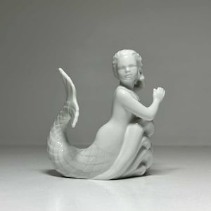 Puede incluir: Figura de sirena de cerámica blanca. La sirena tiene una cola detallada y posa con las manos juntas cerca de la cara. La figura se encuentra sobre un fondo liso, destacando sus delicadas facciones y forma.