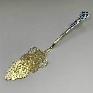Op de afbeelding: Een goudkleurige taartschep met een sierlijk, bloemmotief op het blad. De handgreep heeft een slanke, langwerpige vorm met een decoratief uiteinde met blauwe en gouden accenten. Het wordt waarschijnlijk gebruikt voor het serveren van taarten of gebak.