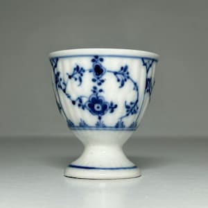 Puede incluir: Taza de huevo de porcelana blanca con un patrón floral azul. La taza tiene una base de pedestal y un borde acampanado. El diseño azul presenta flores y enredaderas estilizadas. La huevera es un diseño clásico, adecuado para servir huevos.