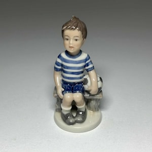 Puede incluir: Figura de porcelana de un niño con una camiseta a rayas azules y blancas y pantalones cortos azules, sentado en un banco. Sostiene un balón de fútbol. La figura está pintada con detalles realistas.