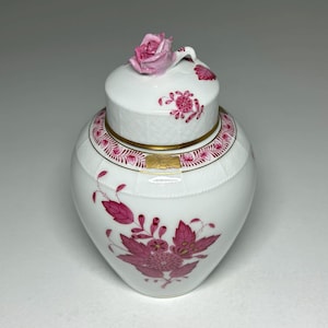May include: A white porcelain jar with a lid, adorned with pink floral designs and a gold band. The lid is topped with a pink rose. The jar's body features a large floral motif, creating an elegant decorative piece.