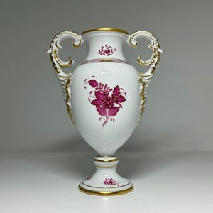 May include: A white porcelain vase with gold detailing and handles. The vase has a floral pattern in pink and gold. The vase stands on a pedestal base and is likely for decorative use.