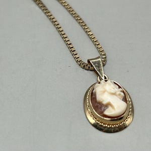 Pode incluir: Um colar de corrente de ouro com um pingente de camafeu que apresenta um perfil esculpido de uma mulher. O camafeu está engastado em uma moldura de ouro com uma borda detalhada.