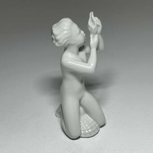 Puede incluir: Una figura de porcelana blanca de una figura arrodillada sosteniendo un objeto en forma de concha. La figura es lisa y detallada, con cabello suelto y una expresión serena. La base es una concha marina.