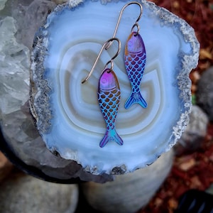 May include: A pair of fish-shaped earrings with a gradient of purple, blue, and gold hues. The earrings have a scale pattern and copper-colored ear wires. The earrings are displayed on a blue and white stone.