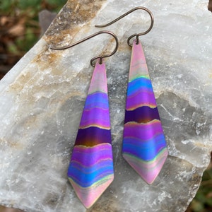 May include: A pair of iridescent purple and blue dangle earrings with a geometric shape. The earrings have a brushed metal finish and are suspended from silver hooks.