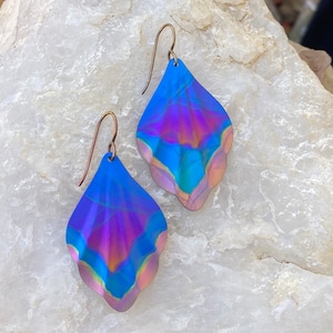 May include: A pair of drop earrings featuring teardrop-shaped metal components with a wavy, scalloped edge. The surface displays an iridescent finish with shifting gradients of vibrant blue, deep purple, and warm copper tones, suspended from simple metal hooks.