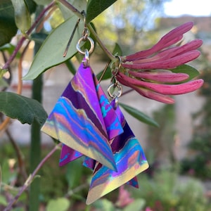 May include: A pair of iridescent triangle earrings with a marbled pattern of purple, blue, and gold. The earrings hang from silver-colored ear wires. The background features green leaves and pink flowers.