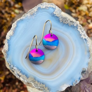 May include: A pair of silver dangle earrings with a round, iridescent blue, pink, and orange painted design. The earrings are displayed on a white agate stone.