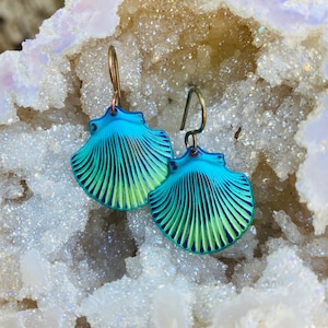 May include: A pair of iridescent blue and green seashell earrings with a bronze wire hanger. The earrings are displayed on a white crystal background.