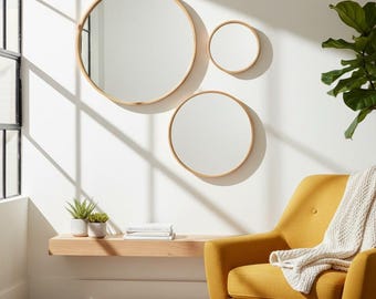 May include: Three circular wall mirrors with light wood frames are mounted on a white wall. Below the mirrors is a floating light wood shelf holding two small potted plants and a stack of books, next to a mustard yellow armchair with a white throw.