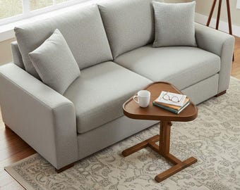 May include: A light gray two-cushion sofa with two matching throw pillows. A small, triangular wooden side table holds a white mug, books, and eyeglasses. The sofa sits on a patterned rug, near a window.