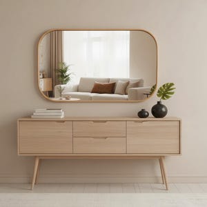 May include: A light wood sideboard with three drawers and two doors, topped with a rounded rectangular mirror. The mirror reflects a living room with a sofa and a window. Decorative items include a black vase and a small black sphere.