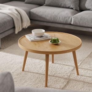 May include: A round, light-colored wooden coffee table with three angled legs. A white bowl, a stack of magazines, and a small green glass object sit on the table. The table is in a living room setting.