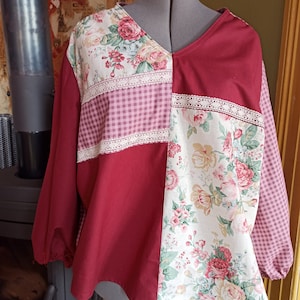 Pode incluir: Uma blusa patchwork com decote em V. Combina tecido bordô liso, estampas florais e xadrez vichy rosa. Acabamentos em renda branca adornam as costuras. As mangas compridas são franzidas nos punhos. A blusa é exibida em um manequim.
