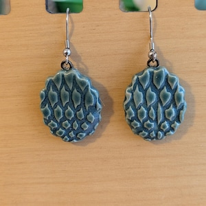 May include: A pair of teal-colored, oval-shaped earrings with a textured, scale-like pattern. The earrings hang from silver-colored ear wires and are displayed against a light wood background.
