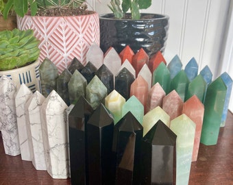 May include: A collection of various crystal towers in shades of white, black, green, red, and blue. The crystals are arranged on a wooden surface, with potted cacti and succulents in the background.