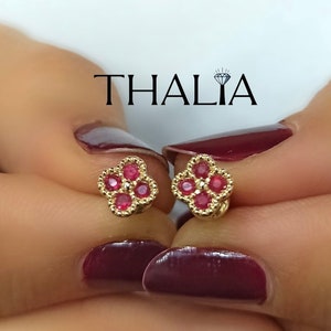 May include: Gold-colored stud earrings, each featuring a four-leaf clover design with a central red gemstone in each leaf. The earrings are held up in front of a background. The word "THALIA" is printed in black.