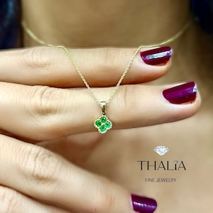 May include: A delicate gold necklace with a small clover pendant set with green gemstones. The pendant is surrounded by a thin gold border. The necklace is displayed on a hand with burgundy nail polish. The image includes the text "THALIA FINE JEWELRY".