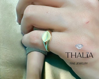 May include: A gold signet ring with a smooth, oval-shaped top. The ring is worn on a person's finger. The image also includes the text "THALIA FINE JEWELRY".